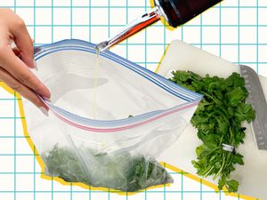 Herb s in a bag being drizzled with olive oil next to a cutting board with a bunch of herbs and a knife.