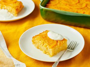 A slice of orange-flavored cake served on a white plate with a dollop of cream and a fork, with the rest of the cake in a green dish in the background