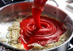 Tomato puree being poured into a pan with sautéed onions on a stovetop surface